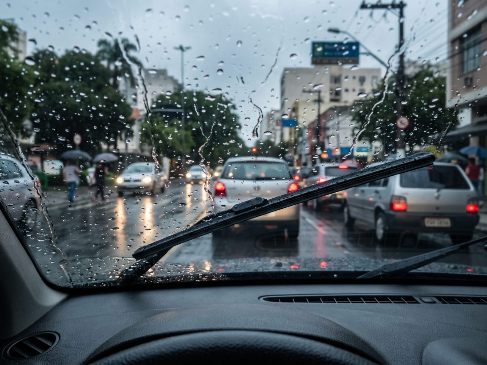 Foto feita de dentro do carro com palheta do limpador de para-brisa em funcionamento durante chuva intensa em via urbana.