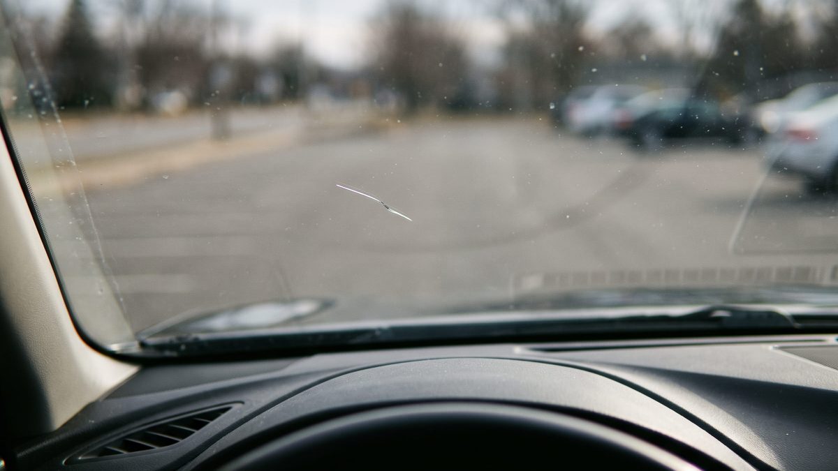 Vista de dentro do carro mostrando um pequeno arranhão no para-brisa, com mão no volante e estacionamento desfocado ao fundo.