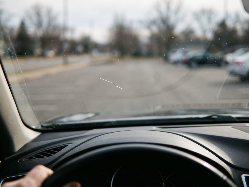 Vista de dentro do carro mostrando um pequeno arranhão no para-brisa, com mão no volante e estacionamento desfocado ao fundo.