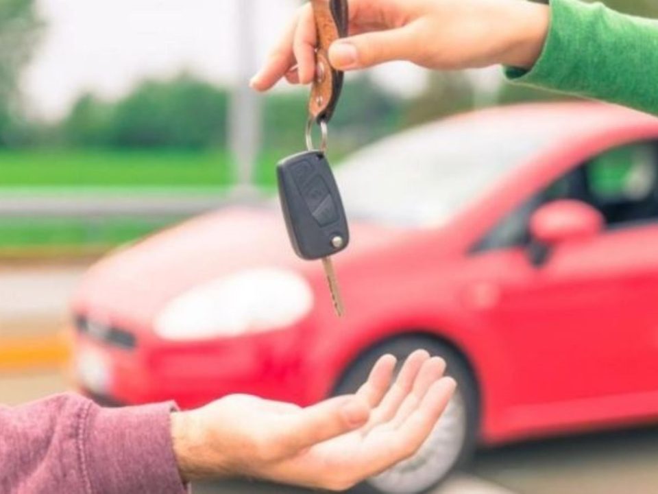 Mão entregando chave de carro para outra pessoa, com carro vermelho desfocado ao fundo, representando a venda de um veículo.