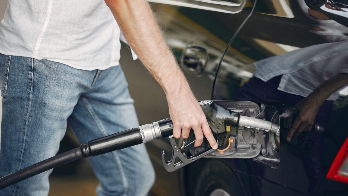 Imagem de uma pessoa abastecendo um carro escuro em um posto de combustível, segurando o bico da bomba de gasolina.