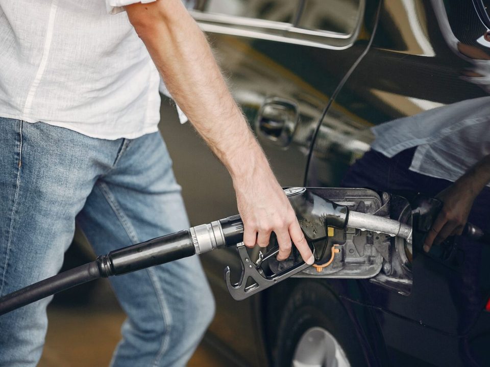 Imagem de uma pessoa abastecendo um carro escuro em um posto de combustível, segurando o bico da bomba de gasolina.