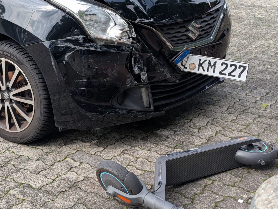 Carro preto com para-choque dianteiro danificado após colisão com patinete elétrico.