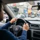 Motorista segurando o volante com as duas mãos enquanto dirige em uma rua movimentada de cidade, com prédios e comércio ao fundo.