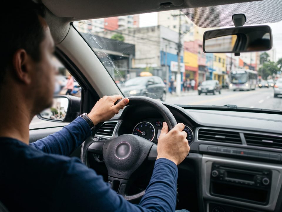 Motorista segurando o volante com as duas mãos enquanto dirige em uma rua movimentada de cidade, com prédios e comércio ao fundo.