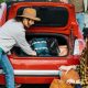Duas mulheres organizando malas e mochilas no porta-malas aberto de um carro vermelho, com um homem ao fundo.