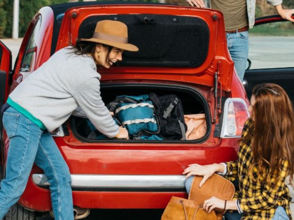 Duas mulheres organizando malas e mochilas no porta-malas aberto de um carro vermelho, com um homem ao fundo.