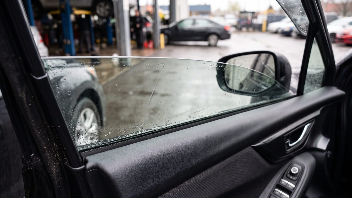 Vidro elétrico do carro levantado até a metade, com gotas de chuva na superfície, visto do interior do veículo em oficina automotiva.