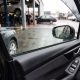 Vidro elétrico do carro levantado até a metade, com gotas de chuva na superfície, visto do interior do veículo em oficina automotiva.