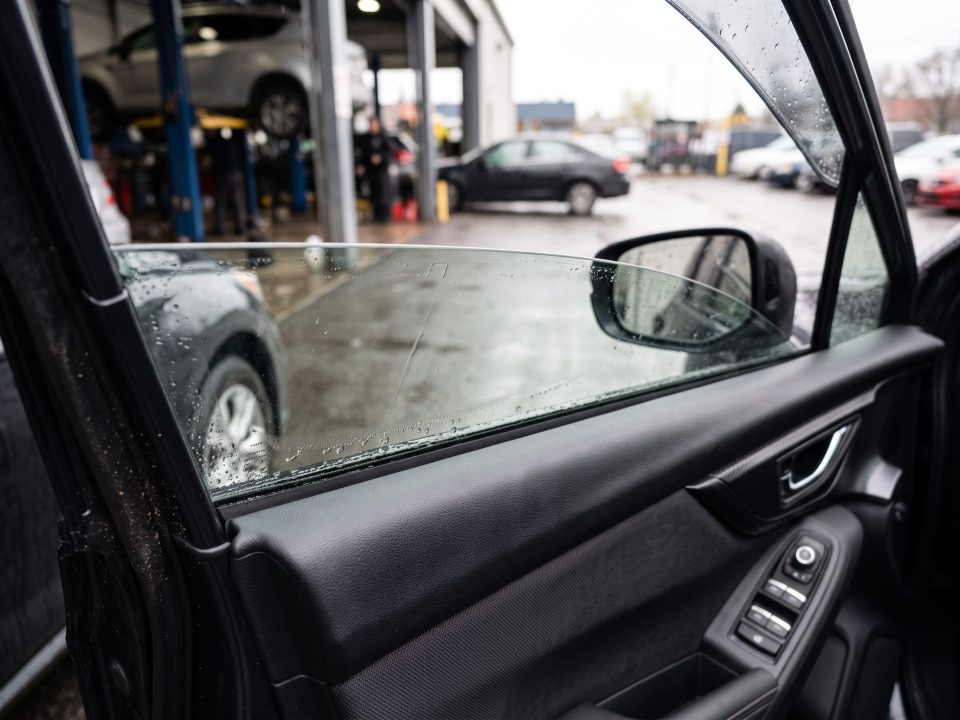 Vidro elétrico do carro levantado até a metade, com gotas de chuva na superfície, visto do interior do veículo em oficina automotiva.