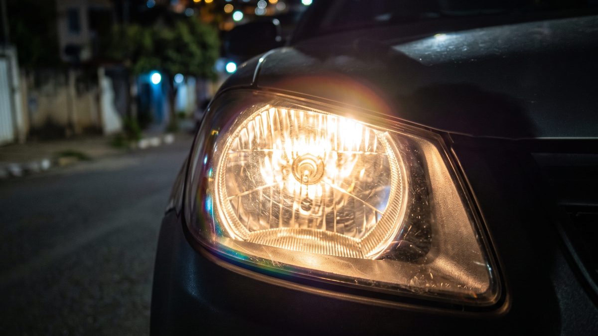 Farol de carro aceso à noite, em destaque, com rua residencial desfocada ao fundo.