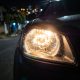 Farol de carro aceso à noite, em destaque, com rua residencial desfocada ao fundo.