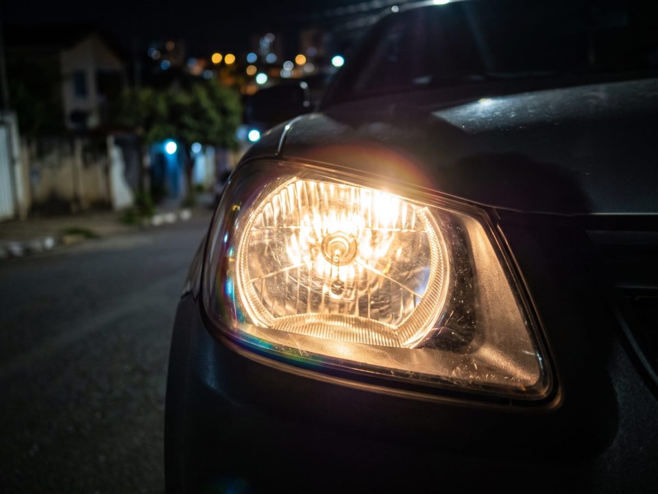 Farol de carro aceso à noite, em destaque, com rua residencial desfocada ao fundo.