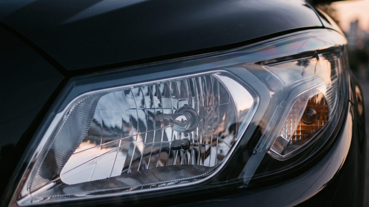 Farol dianteiro de um carro escuro, com lente transparente mostrando o refletor interno e a luz indicadora âmbar ao lado.