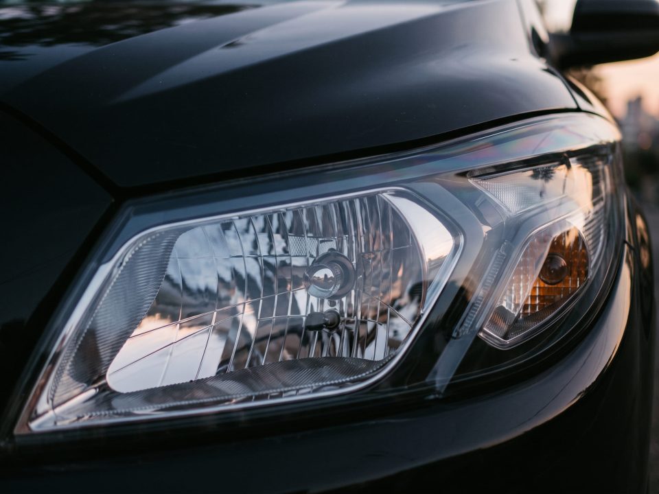 Farol dianteiro de um carro escuro, com lente transparente mostrando o refletor interno e a luz indicadora âmbar ao lado.