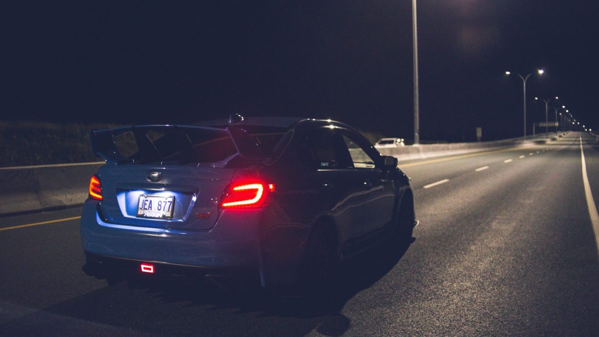 Vista traseira de carro escuro com lanterna de placa iluminada e luz de freio vermelha acesa.