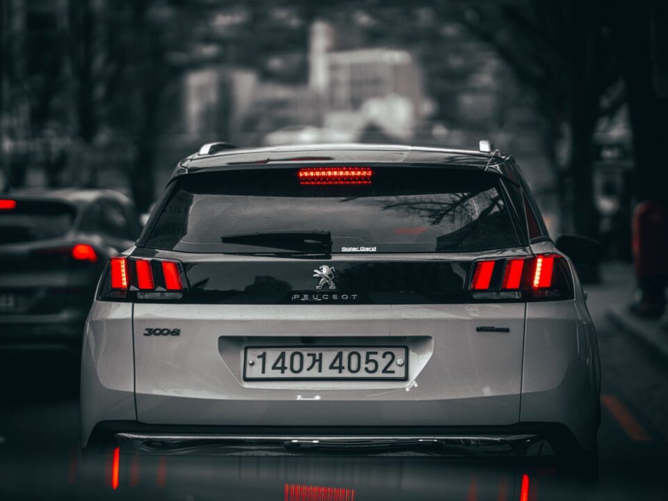 Vista traseira de um Peugeot 3008 branco circulando em via urbana, com lanternas traseiras vermelhas e terceira luz de freio acesas.