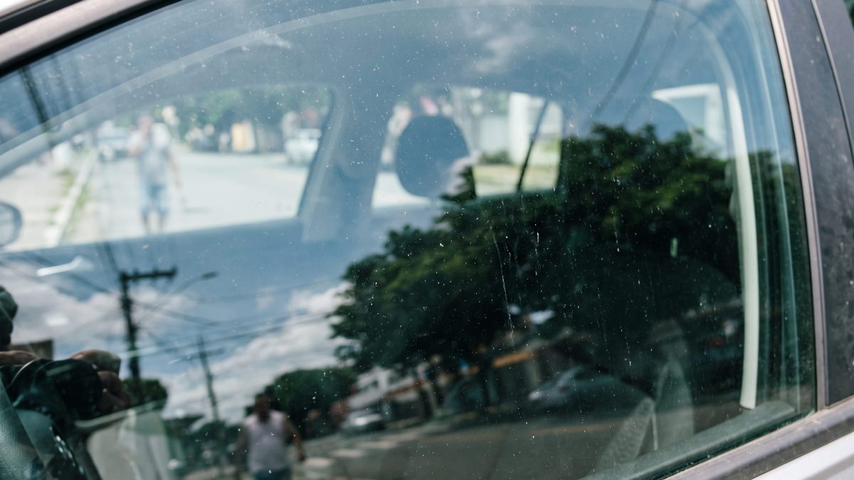 Vidro lateral dianteiro de carro fechado com reflexo de rua urbana e marcas de uso na superfície do vidro.