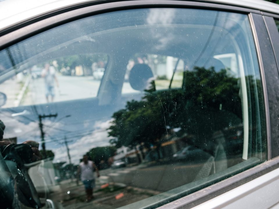 Vidro lateral dianteiro de carro fechado com reflexo de rua urbana e marcas de uso na superfície do vidro.