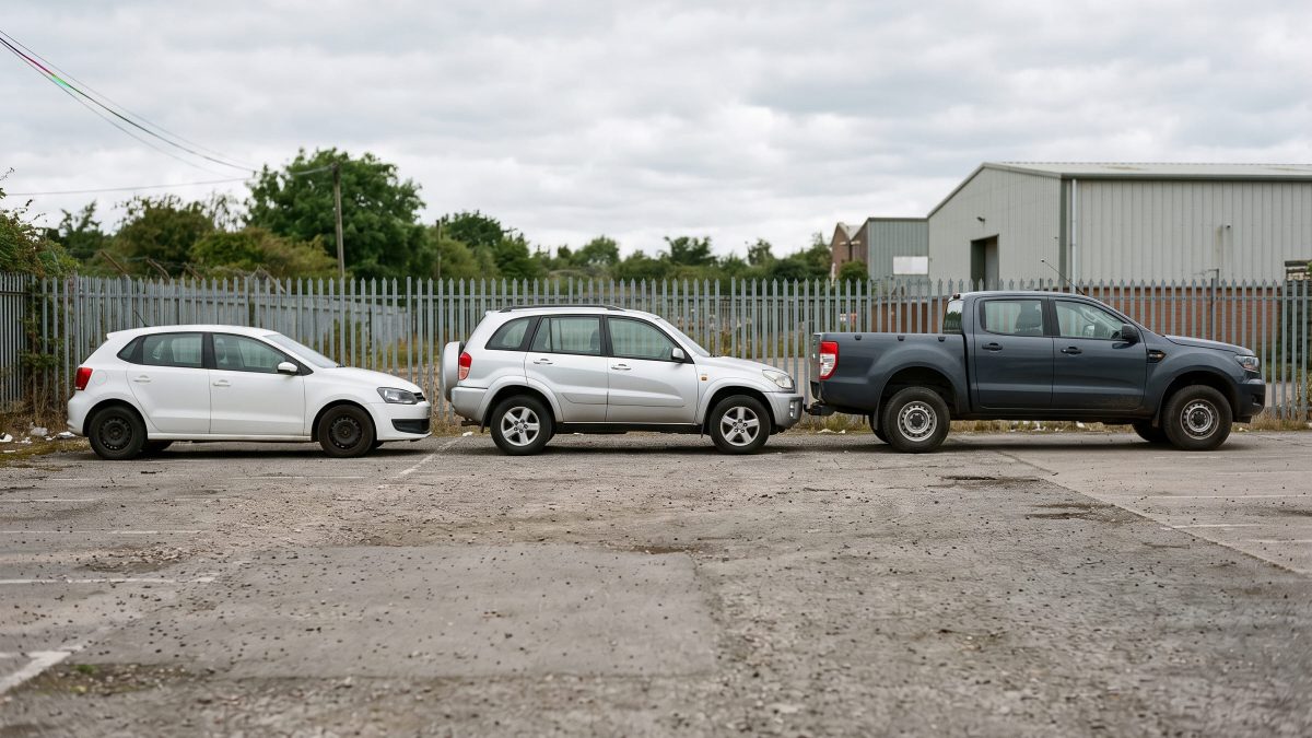 Três carros lado a lado mostrando diferentes tipos de carroceria: hatch branco, SUV prata e picape cinza-escura.