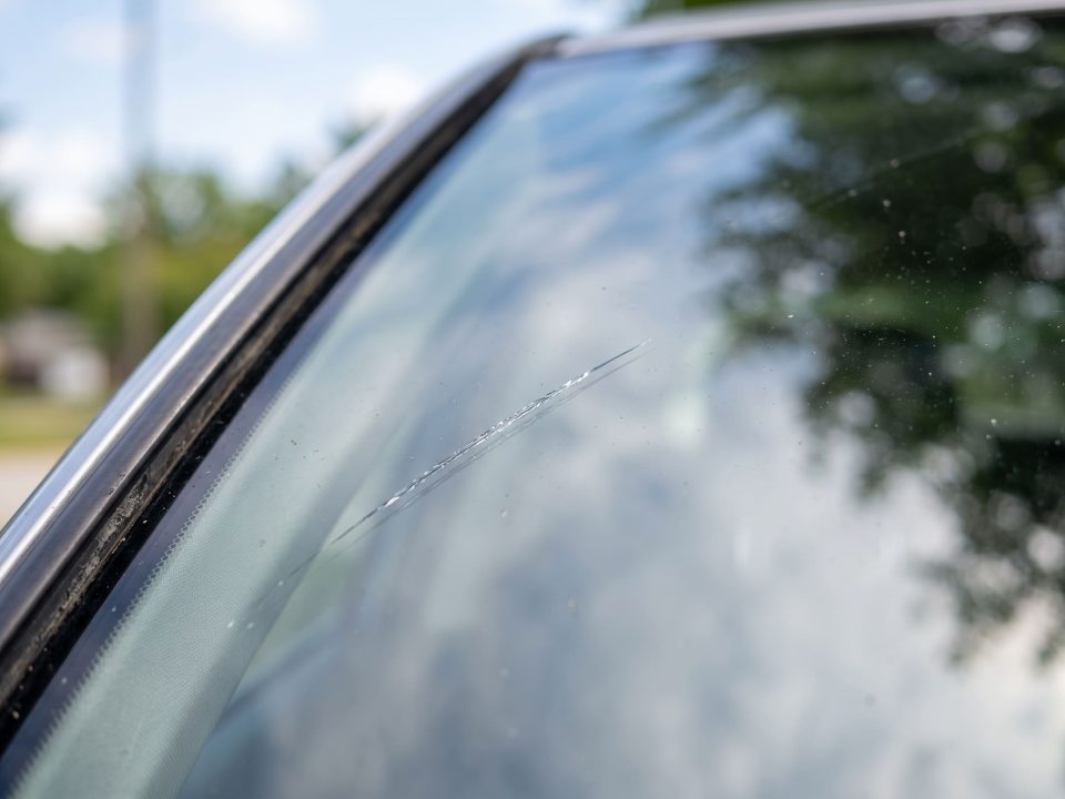 Arranhão fino e superficial bem à esquerda do para-brisa do carro, visto de fora em ângulo lateral.