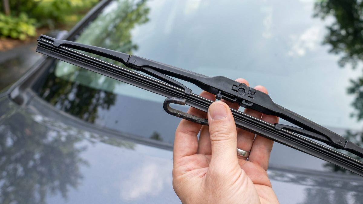 Mão de motorista trocando a palheta do limpador de para-brisa em destaque, com o veículo ao fundo.