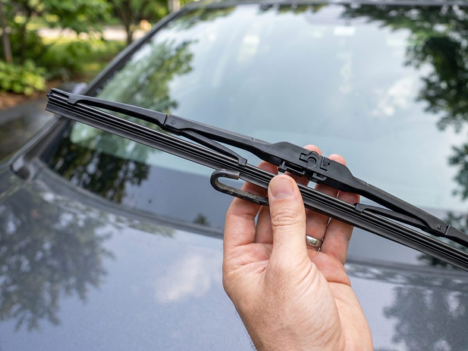 Mão de motorista trocando a palheta do limpador de para-brisa em destaque, com o veículo ao fundo.