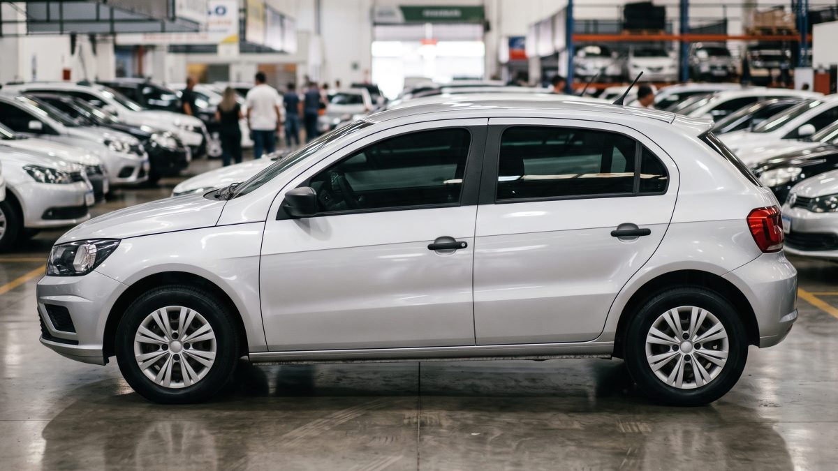 Carro hatch cinza fotografado de perfil dentro de uma loja de veículos seminovos, com outros carros desfocados ao fundo