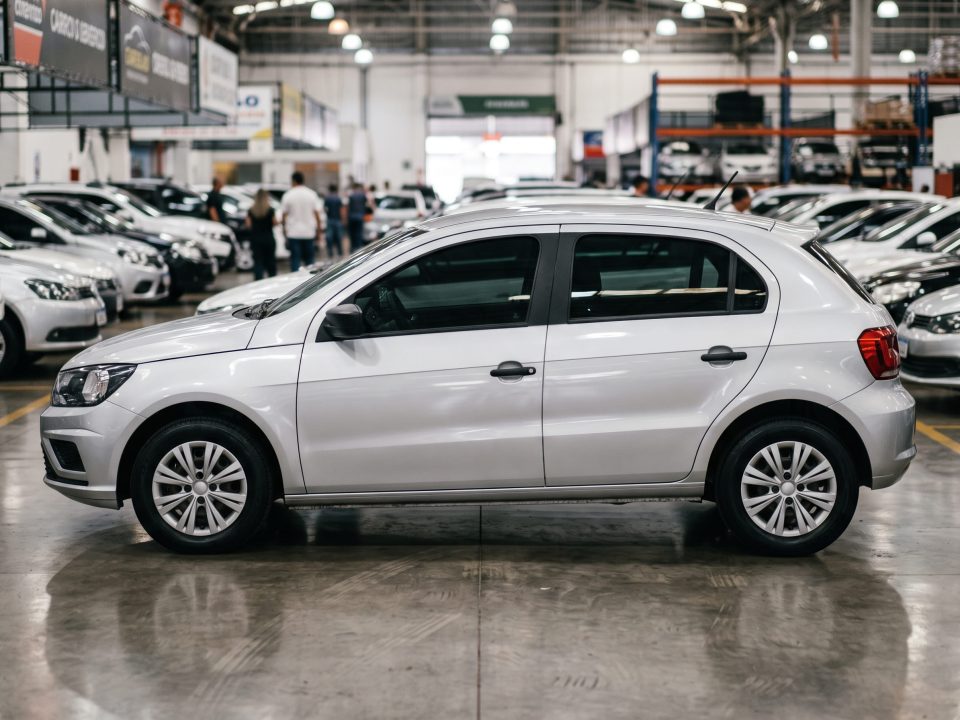 Carro hatch cinza fotografado de perfil dentro de uma loja de veículos seminovos, com outros carros desfocados ao fundo