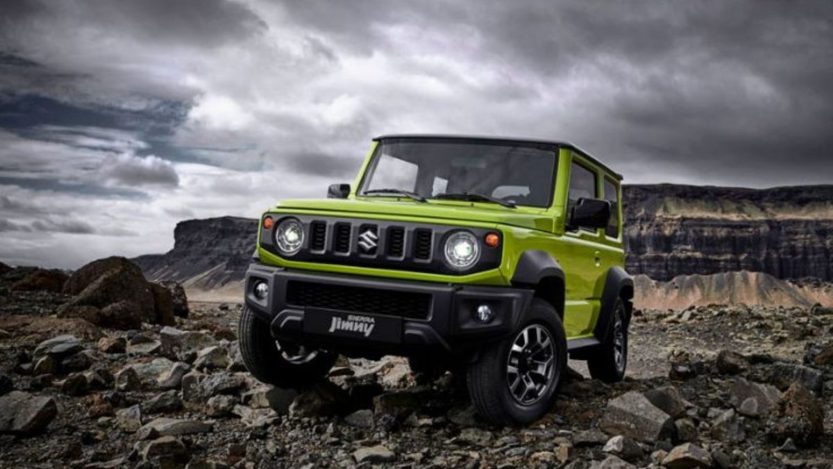 Suzuki Jimny Sierra verde estacionado sobre terreno rochoso, com formações de pedra e céu nublado ao fundo.