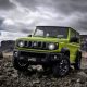 Suzuki Jimny Sierra verde estacionado sobre terreno rochoso, com formações de pedra e céu nublado ao fundo.