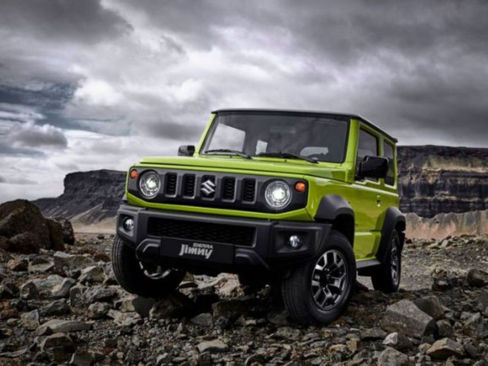 Suzuki Jimny Sierra verde estacionado sobre terreno rochoso, com formações de pedra e céu nublado ao fundo.