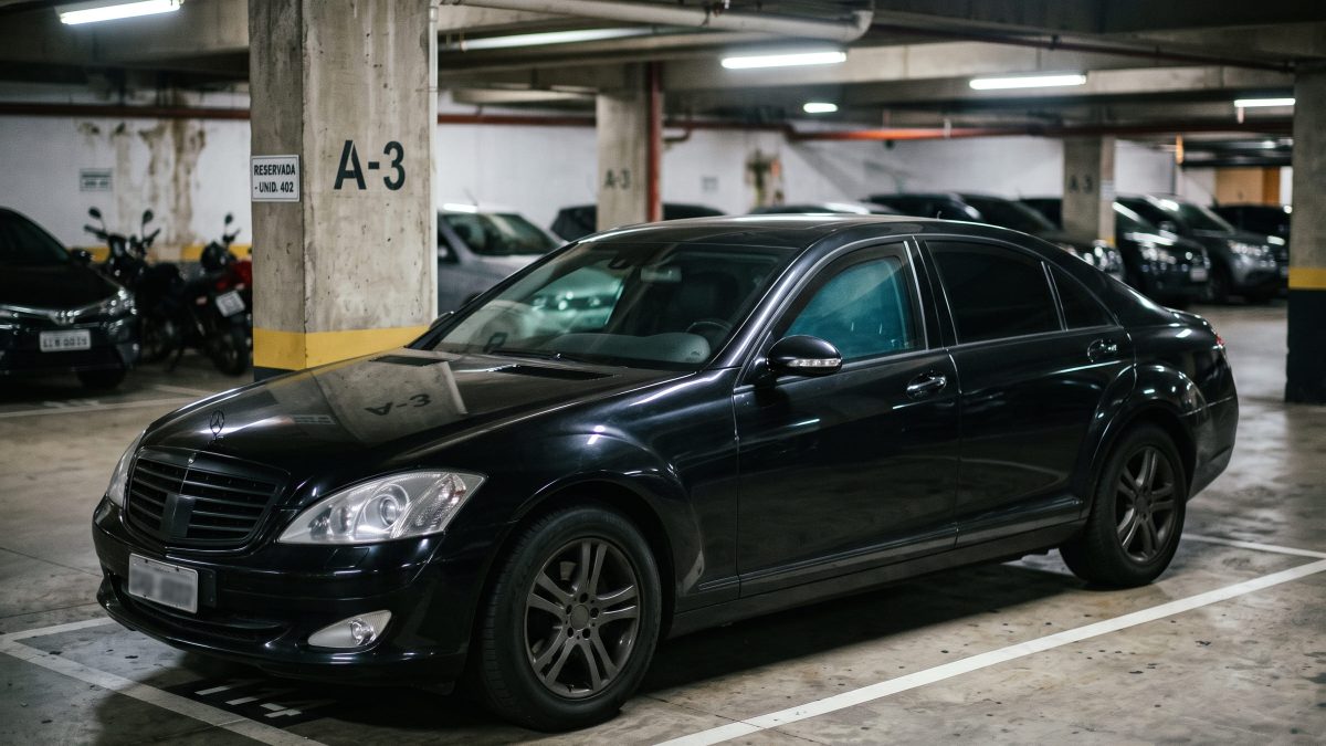 Carro blindado preto estacionado em vaga de garagem coberta de um prédio, com colunas de concreto e outros veículos ao fundo.