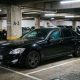 Carro blindado preto estacionado em vaga de garagem coberta de um prédio, com colunas de concreto e outros veículos ao fundo.