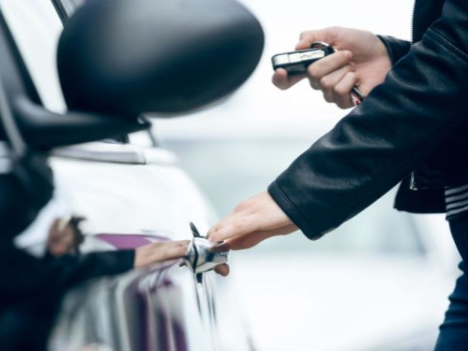 Mão segurando um controle remoto próximo à maçaneta de um carro branco, com o retrovisor visível ao fundo.
