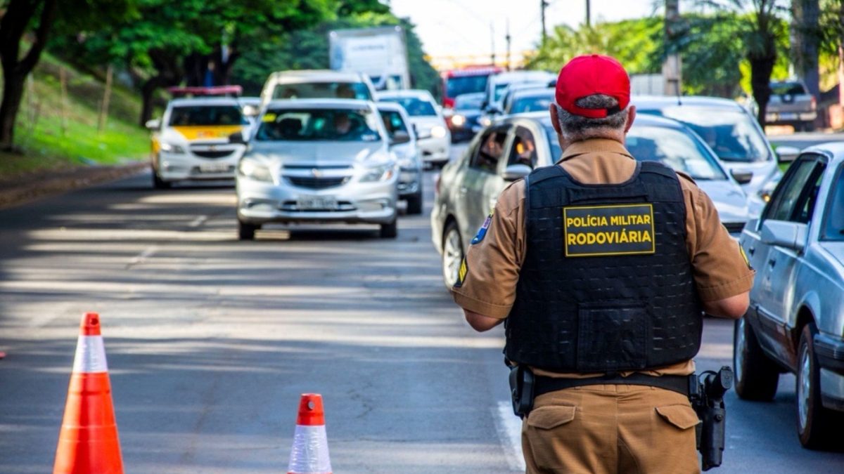 Policial militar rodoviário realiza fiscalização de veículos em uma via com cones de sinalização.