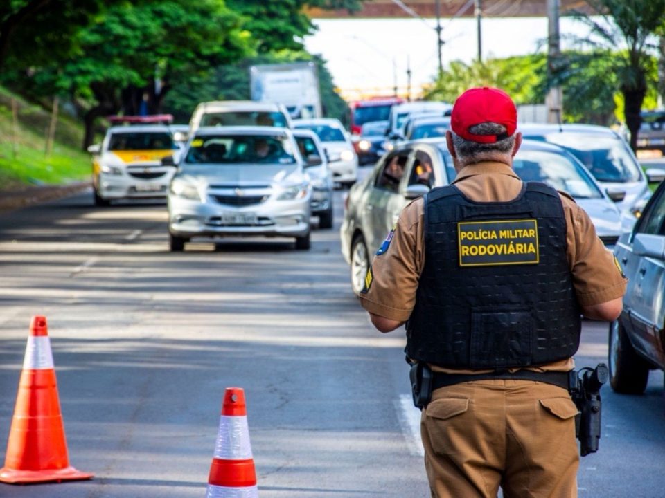 Policial militar rodoviário realiza fiscalização de veículos em uma via com cones de sinalização.