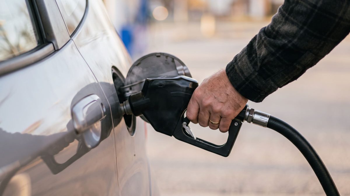 Foto de uma mão abastecendo carro prata com bico de combustível, fundo desfocado de posto de gasolina.