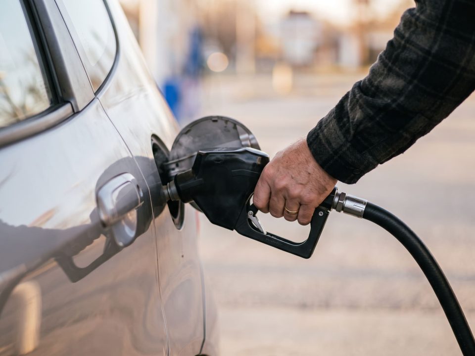 Foto de uma mão abastecendo carro prata com bico de combustível, fundo desfocado de posto de gasolina.