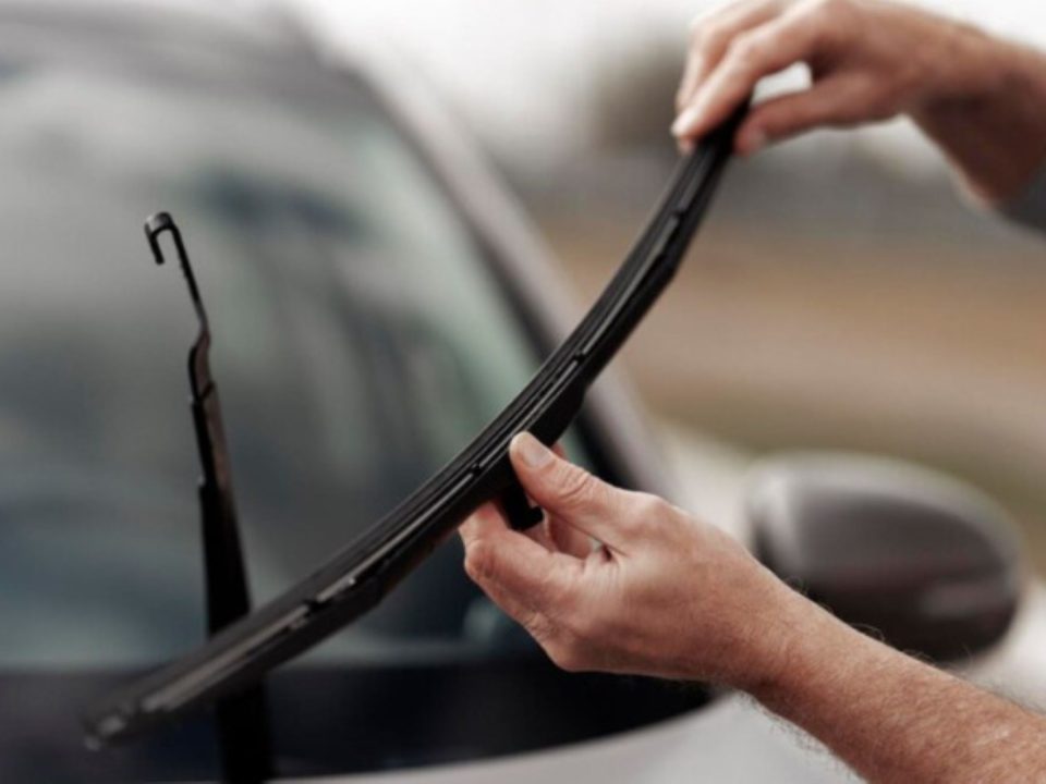 Mãos trocando a palheta do limpador de para-brisa de um carro, com o braço do limpador levantado e a lâmina sendo removida.