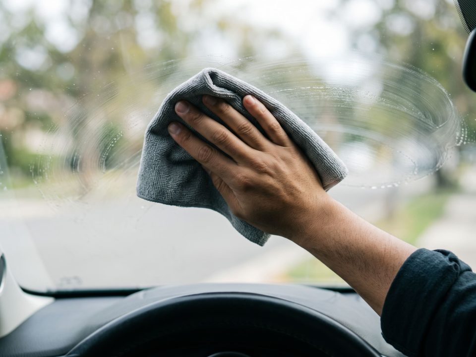 Mão limpando o para-brisa por dentro com pano de microfibra, vista de dentro do carro.