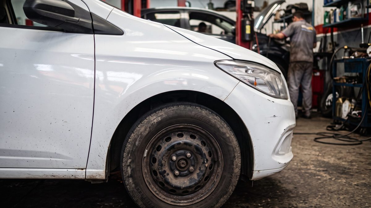 Para-lama e roda dianteira de carro branco em oficina mecânica, com mecânico trabalhando ao fundo.