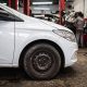 Para-lama e roda dianteira de carro branco em oficina mecânica, com mecânico trabalhando ao fundo.