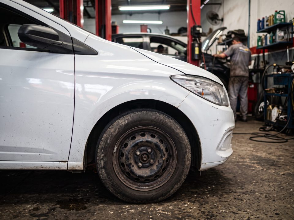 Para-lama e roda dianteira de carro branco em oficina mecânica, com mecânico trabalhando ao fundo.