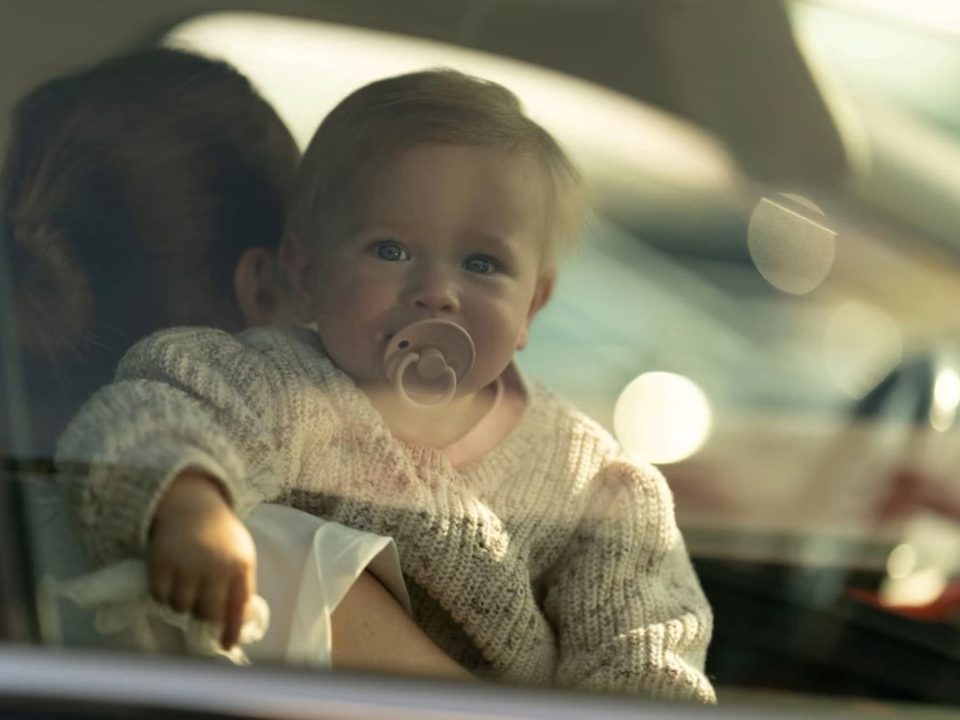 Bebê no colo de um adulto dentro de um carro, visto através do vidro da janela.