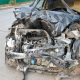 Carro preto com a frente completamente destruída após colisão, motor e estrutura expostos.