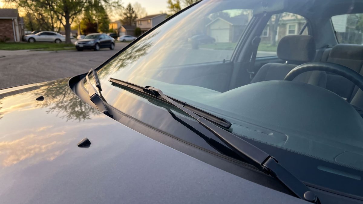 Limpador de para-brisa apoiado sobre o vidro dianteiro (para-brisa) de um carro estacionado numa rua residencial, com a luz do fim de tarde ao fundo.