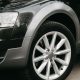 Foto do para-lama e roda de liga leve de um carro preto, com farol dianteiro visível ao fundo.