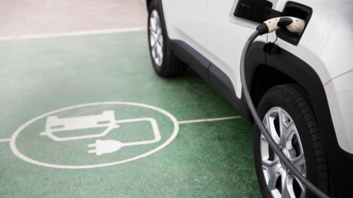 Carro elétrico branco conectado ao cabo de recarga, estacionado sobre símbolo de vaga de carregamento pintado no chão.