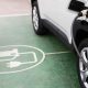 Carro elétrico branco conectado ao cabo de recarga, estacionado sobre símbolo de vaga de carregamento pintado no chão.